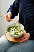 A woman eating a pho bowl with bok choy, radish and rice noodles