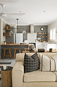 Scatter cushions on pale sofa bed, tree stump side table and kitchen with counter and grey wall in background in open-plan interior