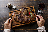 Grilled T-bone steak on serving board, fork and knife in male hands and red wine on wooden table background