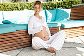 A pregnant woman wearing a white outfit sitting cross-legged on a terrace