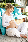 A pregnant woman wearing a white outfit sitting cross-legged on a terrace