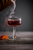Red cocktail in vintage coupe glass with smoke pouring in on a rustic, gray wood surface