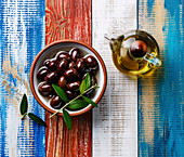 Kalamata-Oliven und Olivenöl in Glaskaraffe auf Holzuntergrund (Aufsicht)