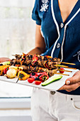 A woman serving lamb shish kebabs with grilled peppers, shallots, fresh cherry tomatoes and cucumber