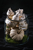 Halloween: jar with white meringue ghosts with chocolate eyes, decor skulls, moss and pumpkin over black background