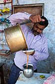 Mann giesst Tee aus Messingtopf durch Sieb in Zinngefäß, Dorfmarkt in Diggi, Rajasthan, Indien