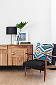 Table lamp and large leaf in vase on top of sideboard with patchwork wooden front next to patterned cushion on armchair