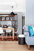 Colourful scatter cushions on grey sofa and side table in front of dining area with classic chairs and crockery on open shelves