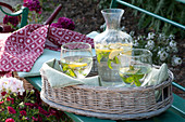 Wicker tray with lemon-mint water in bottle and glasses on bench
