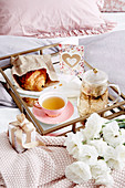 Breakfast tray with tea, croissant and greeting card on bed, flowers and small gift