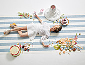 A woman lying on the floor with a paintbrush in her hand, surrounded by doughnuts and cakes