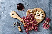 Wooden serving board with fresh red grapes, walnuts, goat and cheddar cheese, with cheese knife and glass of red wine