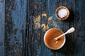 Glass jar of homemade salted caramel sauce with spoon, brown sugar and bowl of salt