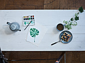 Teekanne, Farbkasten, gemaltes Pflanzenblatt, Tee und Kuchen