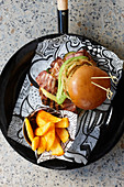 A burger with avocado and bacon, served with french fries in a small metal container