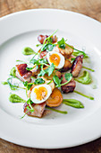 Pan fried bacon and scallops with quail eggs and a pea puree with pea shoots on a white plate and wooden table