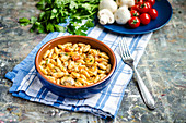 Tomato and mushroom fresh homemade pasta in a terracotta bowl on a blue and white towel, a fork and ingredients and herbs in the background