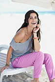 A young, long-haired woman on a lounger on a beach wearing a grey blouse and pink trousers