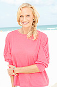 A blonde woman by the sea wearing a pink blouse