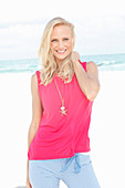 A blonde woman by the sea wearing a pink top and checked trousers