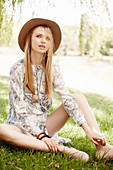 Junge blonde Frau mit Hut im Kleid auf der Wiese sitzend