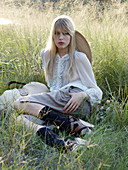 A young blonde woman sitting in a meadow wearing a ruffled blouse, shorts, socks and sandals