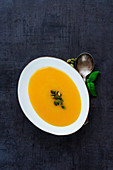 Traditional pumpkin cream soup in white bowl with fresh green basil leaves, spices and pumpkin seeds on dark vintage stone background