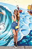 Junge Frau mit Sonnenbrille in grünem Badeanzug und kurzer Jeans-Latzhose vor Graffiti
