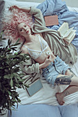 A young woman with pink hair laying on bed wearing a bra and shorts