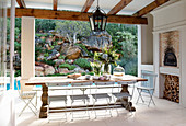 Long oak table and folding chairs on roofed terrace with tiled floor and view of planted rock face