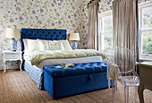 Double bed with royal-blue headboard and ottoman in bedroom with floral wallpaper