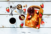 Rural breakfast with fresh croissant, cup of coffee, fruity jam, honey and strawberries on rustic white wooden background