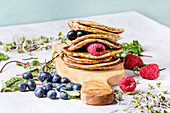 Stack of vegan chickpea pancakes on olive wood cutting board with green salad, sprouts, berries, over grey kitchen table