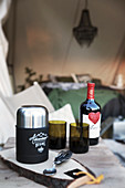 Besteck, Thermoskanne, Gläser und eine Flasche Rotwein auf Holztisch vor Zelt
