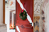 Winterlich dekorierter Eingangsbereich mit Gesteck in Fensternische und Türkranz an roter Holztür