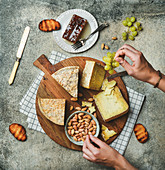 Flat-lay of cheese platter with cheese assortment, green grapes, bread, honey and nuts with female hands reaching to food