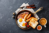English breakfast in cooking pan with fried egg, sausage, bacon, beans, toast and coffee on dark background