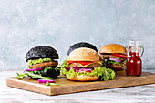 Set of homemade burgers in black and white buns with avocado, tomato sauce, lettuce, arugula, cheese, onion on wood serving board