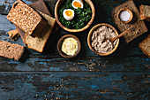 Variety loaves of sliced homemade rye bread whole grain and seeds for breakfast with olive wood bowls of butter, liver paste, salt, spinach, eggs