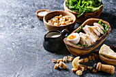 Bowls of ingredients for cooking classic Caesar salad: Sliced baked chicken breast, green roman salad, parmesan cheese, egg, croutons, salt, jug of sauce