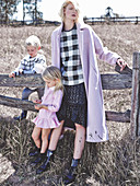 A blonde woman wearing a black-and-white checked blouse, a skirt and a lilac coat with two children in front of a paddock