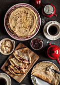 Plates with crepes with jam jar, coffee cup, red coffee pot and red sugar bowl on dark background