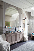 Mirror on wall above ornaments on console cabinet flanked by loose-covered chairs