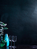 Weingläser, blaue Vase und grüne Glaskaraffe vor dunklem Hintergrund
