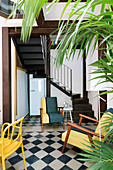Farbenfrohe Sitzmöbel auf Schachbrettmusterboden und Grünpflanzen im Wohnraum mit Treppenaufgang