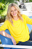 A young blonde woman wearing a yellow jumper sitting outside on a upholstered bench