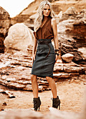 A blonde woman wearing a brown top and a grey skirt standing in front of rocks