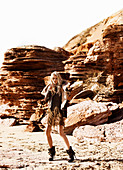A blonde woman wearing a leather jacket and a brown mini dress standing in front of rocks