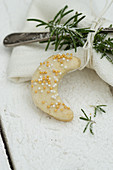 A crescent-shaped biscuit with sugar sprinkles next to a napkin with a silver fork and a sprig of rosemary