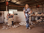 A young woman in a barn wearing a t-shirt and a matching long skirt with a man shearing sheep in the background